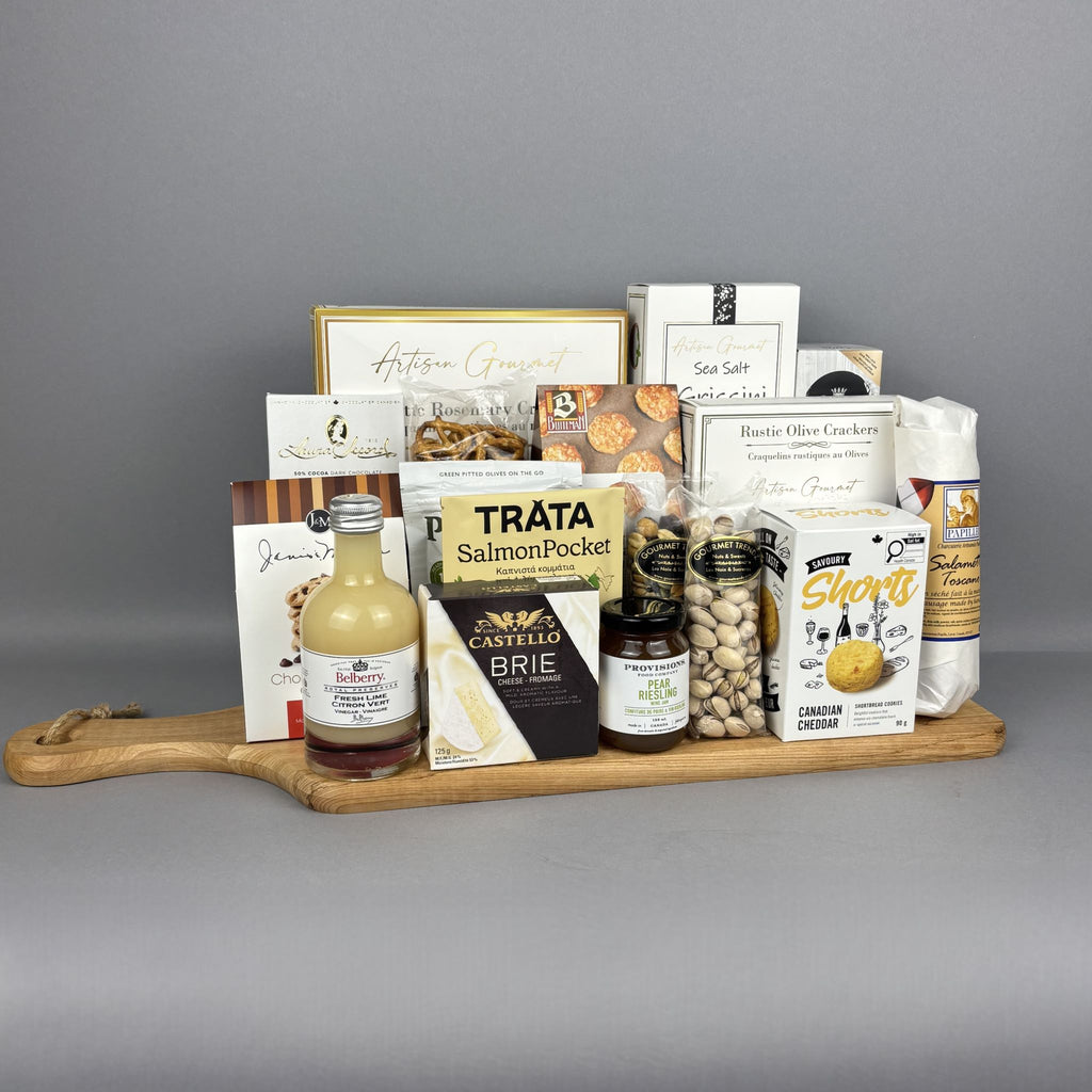 A gourmet gift hamper arranged on a wooden serving board. The assortment includes artisanal cheeses, crackers, nuts, a bottle of fresh lime vinegar, and packaged snacks from brands such as Provisions, Castello Brie, and Belberry.