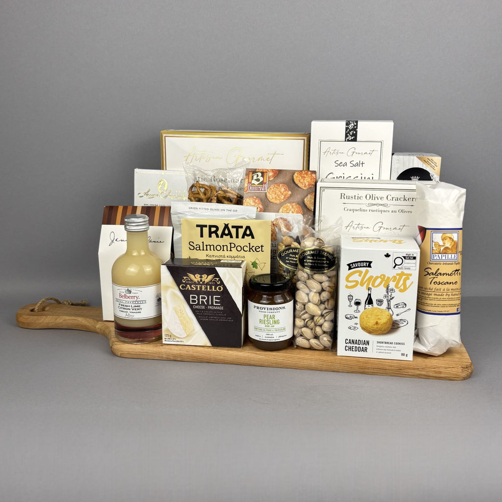 A gourmet gift hamper arranged on a wooden serving board. The assortment includes Tráta Salmon Pocket crackers, Castello Brie cheese, Shorts biscuits, breadsticks and crackers, pistachios, and a bottle of vinegar.