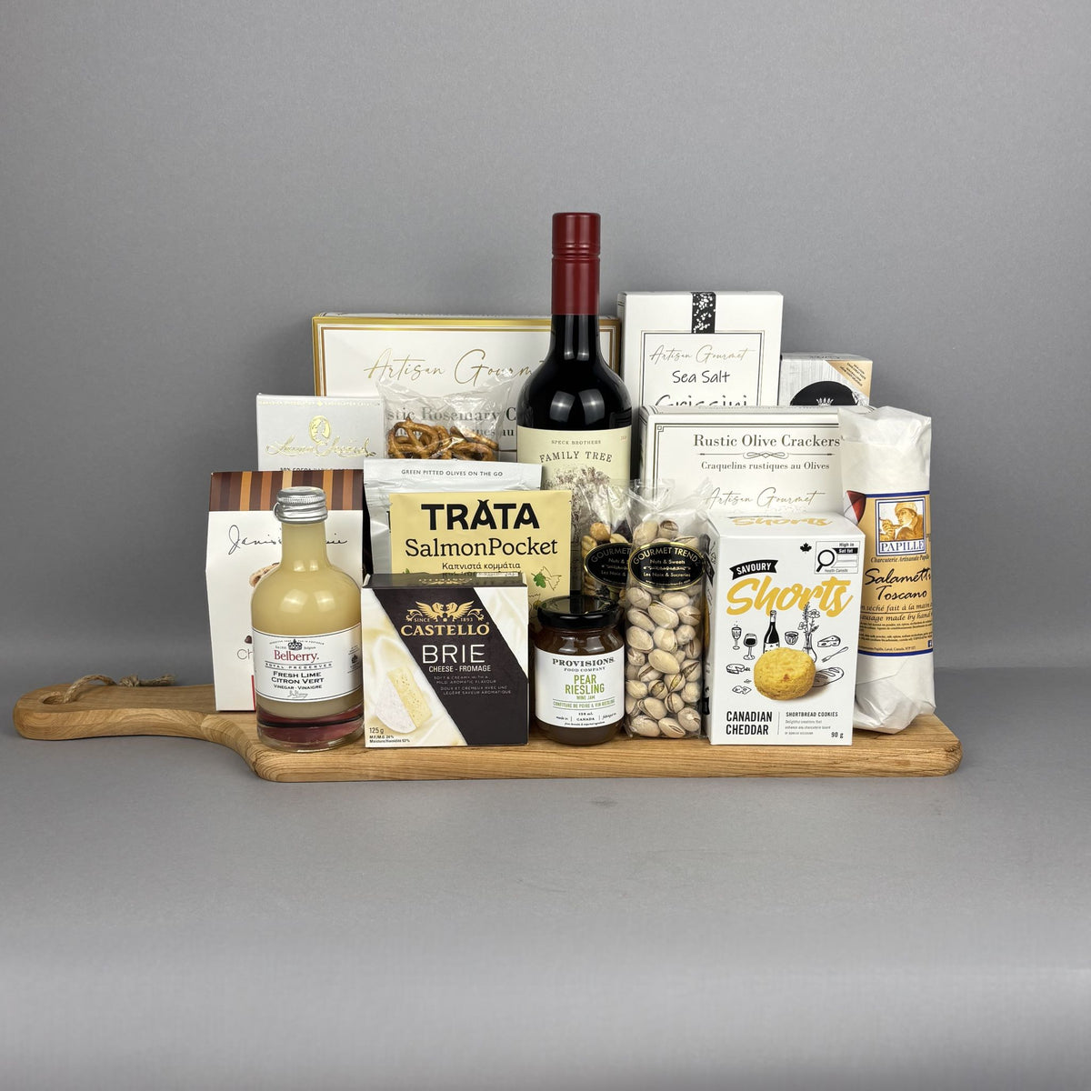 A gourmet gift hamper arranged on a wooden serving board. The hamper contains a bottle of red wine, a bottle of fresh lime vinegar, a box of brie cheese, a box of Shorts Canadian cheddar crackers, a bag of pistachios, and several sweet and savoury items.