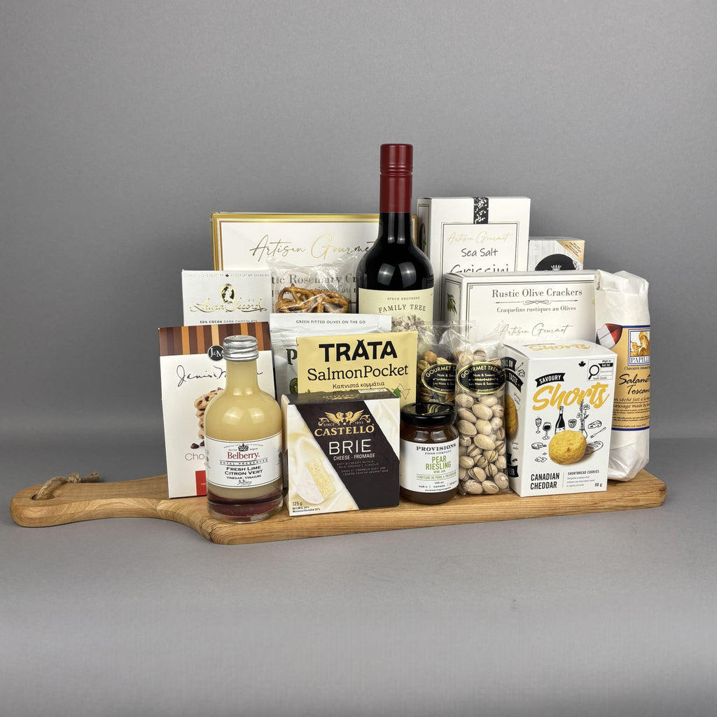 A gourmet gift hamper arranged on a wooden cutting board. The hamper includes a bottle of red wine, salmon pocket, crackers, brie cheese, pistachios, sea salt breadsticks, rustic olive crackers, and other packaged food items.