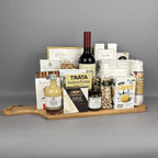 A gourmet gift hamper arranged on a wooden cutting board. The hamper includes a bottle of red wine, salmon pocket, crackers, brie cheese, pistachios, sea salt breadsticks, rustic olive crackers, and other packaged food items.