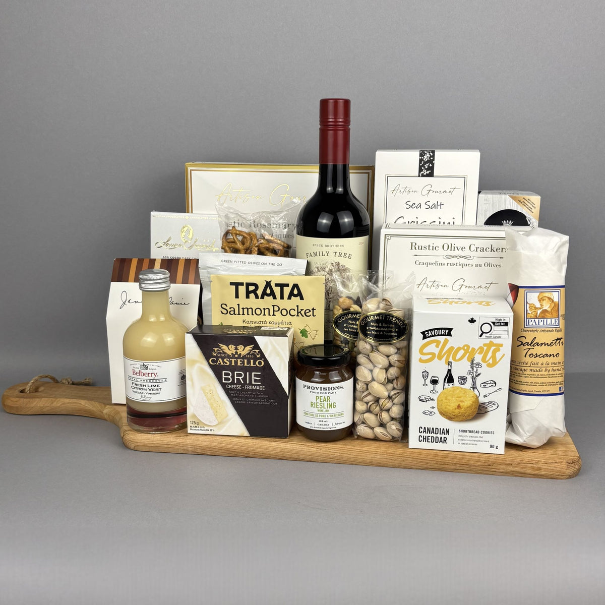 A gourmet gift hamper arranged on a wooden serving board. The hamper includes a bottle of red wine, a small bottle of fresh lime vinegar, a box of Castello Brie, Tráta Salmon Pocket, crackers, Provisions jam, Sea Salt breadsticks, and various packaged snacks and cheeses.