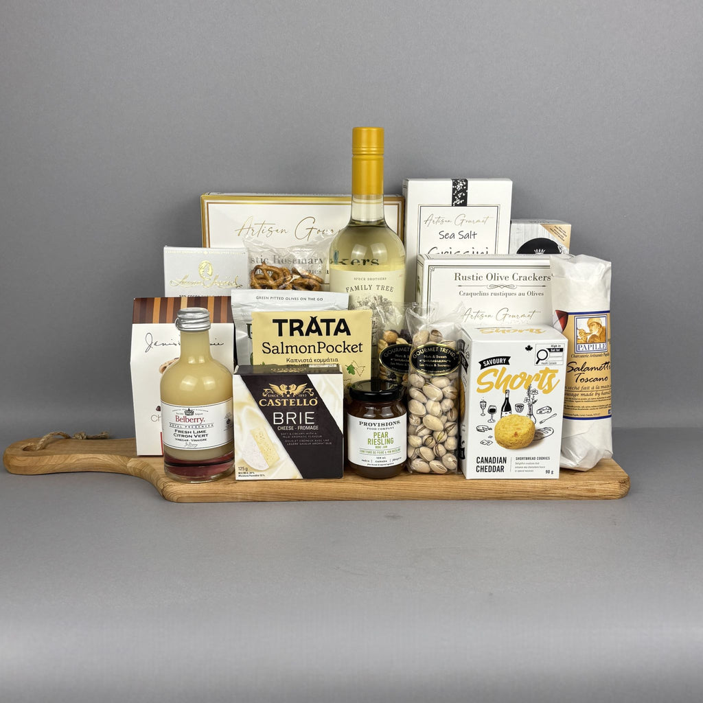 A gourmet gift hamper arranged on a wooden serving board. The hamper includes a bottle of white wine, a small bottle of vinegar, a box of Brie cheese, Tráta Salmon Pocket, Castello Brie cheese, Short's Canadian Cheddar crackers, a bag of pistachios, and several other packaged snacks and treats.