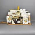 A gourmet gift hamper arranged on a wooden serving board. The hamper includes a bottle of white wine, a small bottle of vinegar, a box of Brie cheese, Tráta Salmon Pocket, Castello Brie cheese, Short's Canadian Cheddar crackers, a bag of pistachios, and several other packaged snacks and treats.