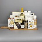 A gourmet gift hamper arranged on a wooden serving board. The hamper includes a bottle of white wine, a small bottle of vinegar, a box of Brie cheese, Tráta Salmon Pocket, Castello Brie cheese, Short's Canadian Cheddar crackers, a bag of pistachios, and several other packaged snacks and treats.