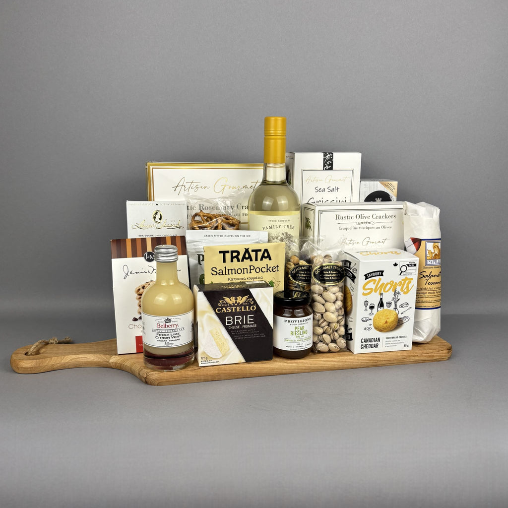 A gourmet gift hamper arranged on a wooden serving board. The assortment includes a bottle of white wine, a bottle of fresh lime vinegar, Provisions jam,  crackers, Brie cheese, pistachios, cookies, and other packaged snacks and sweets.
