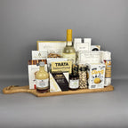 A gourmet gift hamper arranged on a wooden serving board. The assortment includes a bottle of white wine, a bottle of fresh lime vinegar, Provisions jam,  crackers, Brie cheese, pistachios, cookies, and other packaged snacks and sweets.