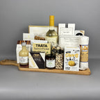 A gourmet gift hamper arranged on a wooden serving board. The board holds a bottle of white wine, a bottle of vinegar, a salmon pocket pouch, brie cheese, pistachios, crackers, and other packaged food items.
