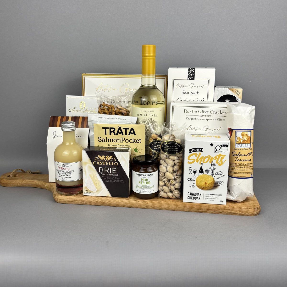 A gourmet gift hamper arranged on a wooden serving board. The board holds a bottle of white wine, a bottle of vinegar, a salmon pocket pouch, brie cheese, pistachios, crackers, and other packaged food items.