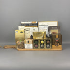 A gourmet chocolate gift board arranged on a light wood cutting board. The assortment includes boxes of Sprucewood cookies and Cantucci biscotti, Allessia chocolate chip biscotti, Made toffee truffles, and a bag of Lindt Lindor truffles.