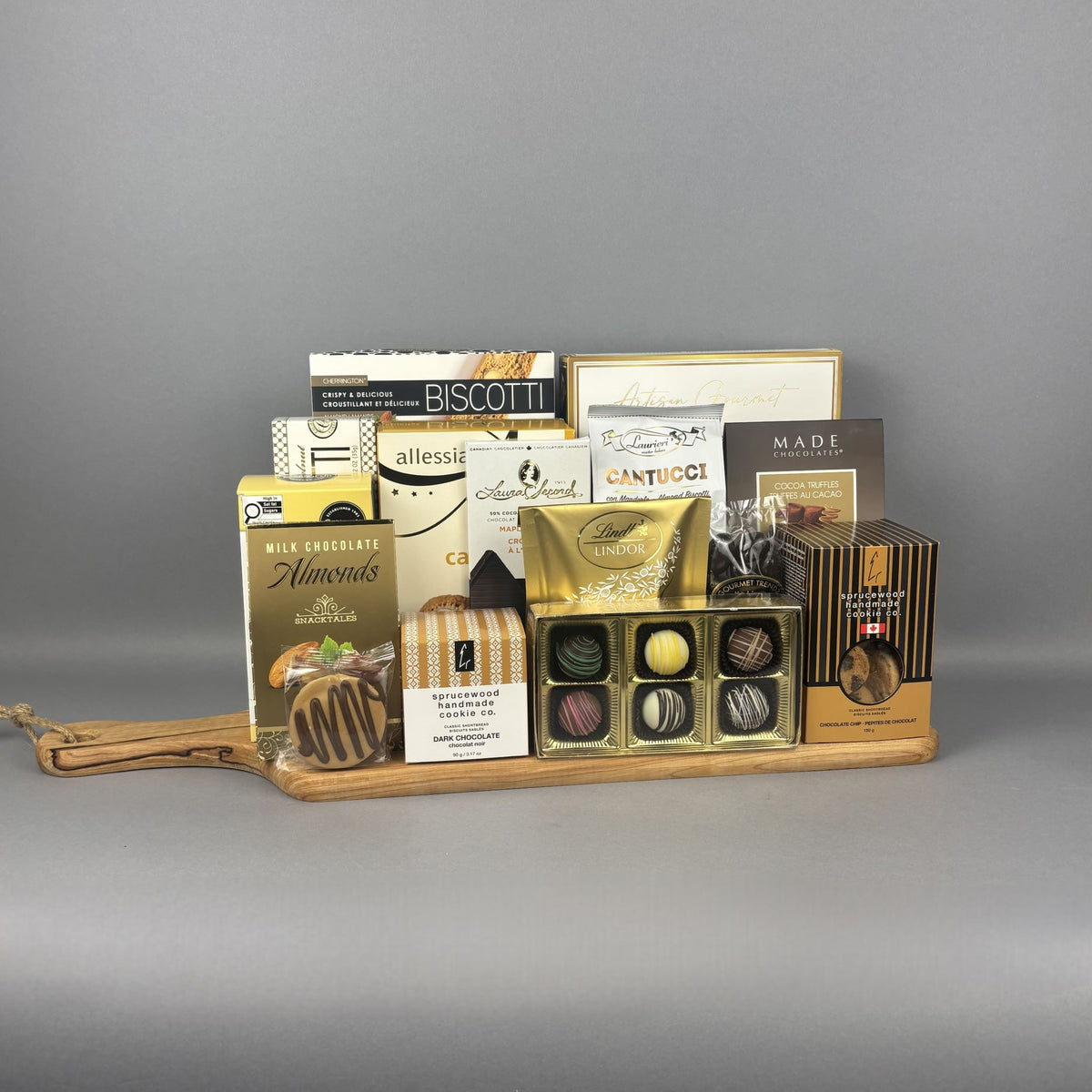 A gourmet chocolate and snack gift board arranged on a wooden serving tray. The assortment includes boxes of Biscotti, Allessia cookies, Lindt Lindor chocolates, and other sweet treats in elegant packaging.