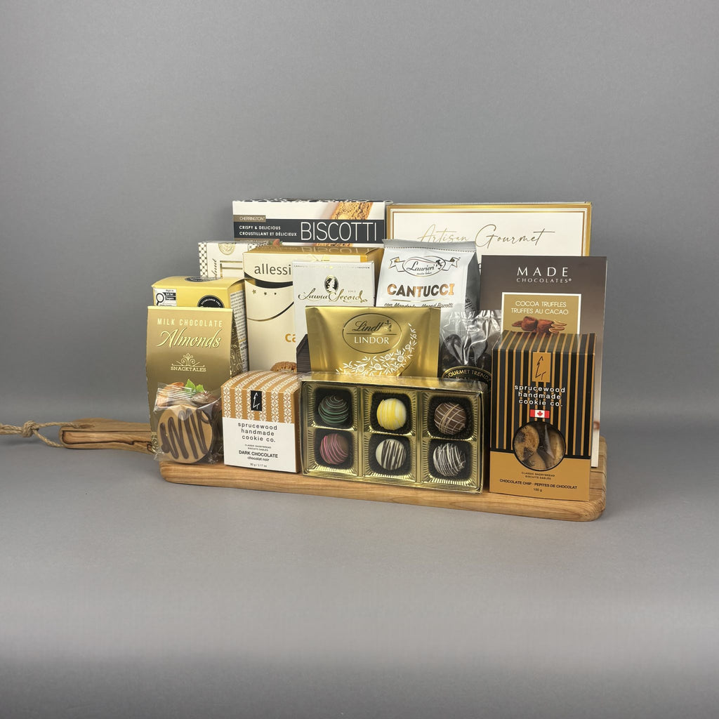 A gourmet chocolate gift tray arranged on a light wood cutting board. The assortment includes boxes of biscotti, chocolates, and sweets from brands such as Sprucewood, Lindt, Laura Secord, and Cookie It Up.