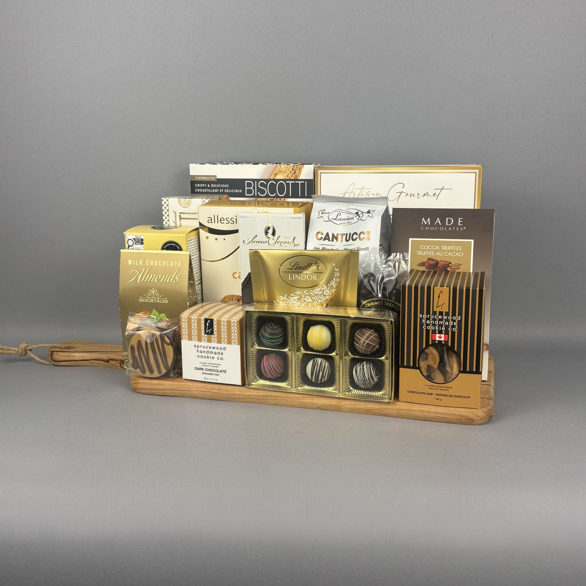 A gourmet chocolate gift tray arranged on a light wood cutting board. The assortment includes boxes of biscotti, chocolates, and sweets from brands such as Sprucewood, Lindt, Laura Secord, and Cookie It Up.