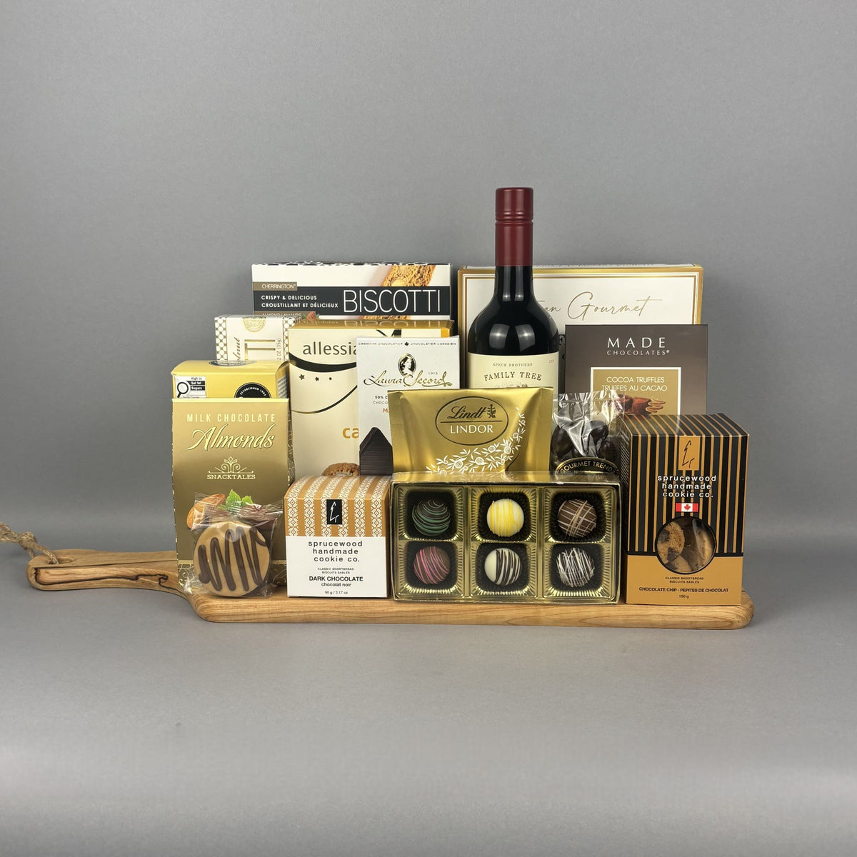 A gourmet gift hamper arranged on a wooden tray, featuring a bottle of Cabernet-Merlot wine, assorted chocolates, biscuits, almonds, and packaged treats.