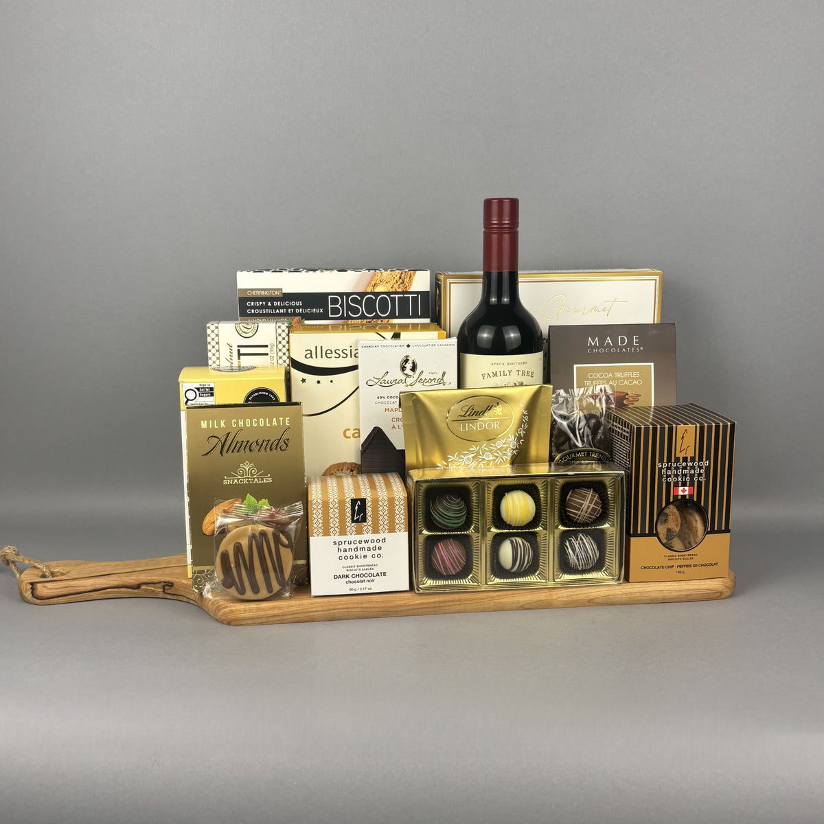 A gourmet gift hamper arranged on a wooden tray, featuring a bottle of red wine, assorted chocolates, biscuits, and packaged treats.