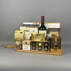 A gourmet gift hamper featuring an assortment of chocolates, biscuits, a bottle of red wine, and packaged treats arranged on a wooden tray.