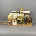 A luxury gourmet gift hamper arranged on a wooden tray. The hamper includes a bottle of white wine, assorted chocolates in a gold tin, biscotti, almonds, and other packaged treats.