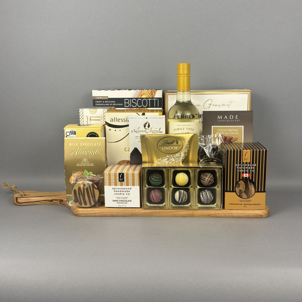 A luxury gourmet gift hamper arranged on a wooden tray. The hamper includes a bottle of white wine, assorted chocolates in a gold tin, biscotti, almonds, and other packaged treats.