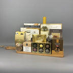 A gourmet gift hamper arranged on a light wood tray. The hamper includes a bottle of white wine, assorted biscotti, chocolates, truffles, and packaged snacks from brands such as Sprucewood, Laura Secord, and Lindt.