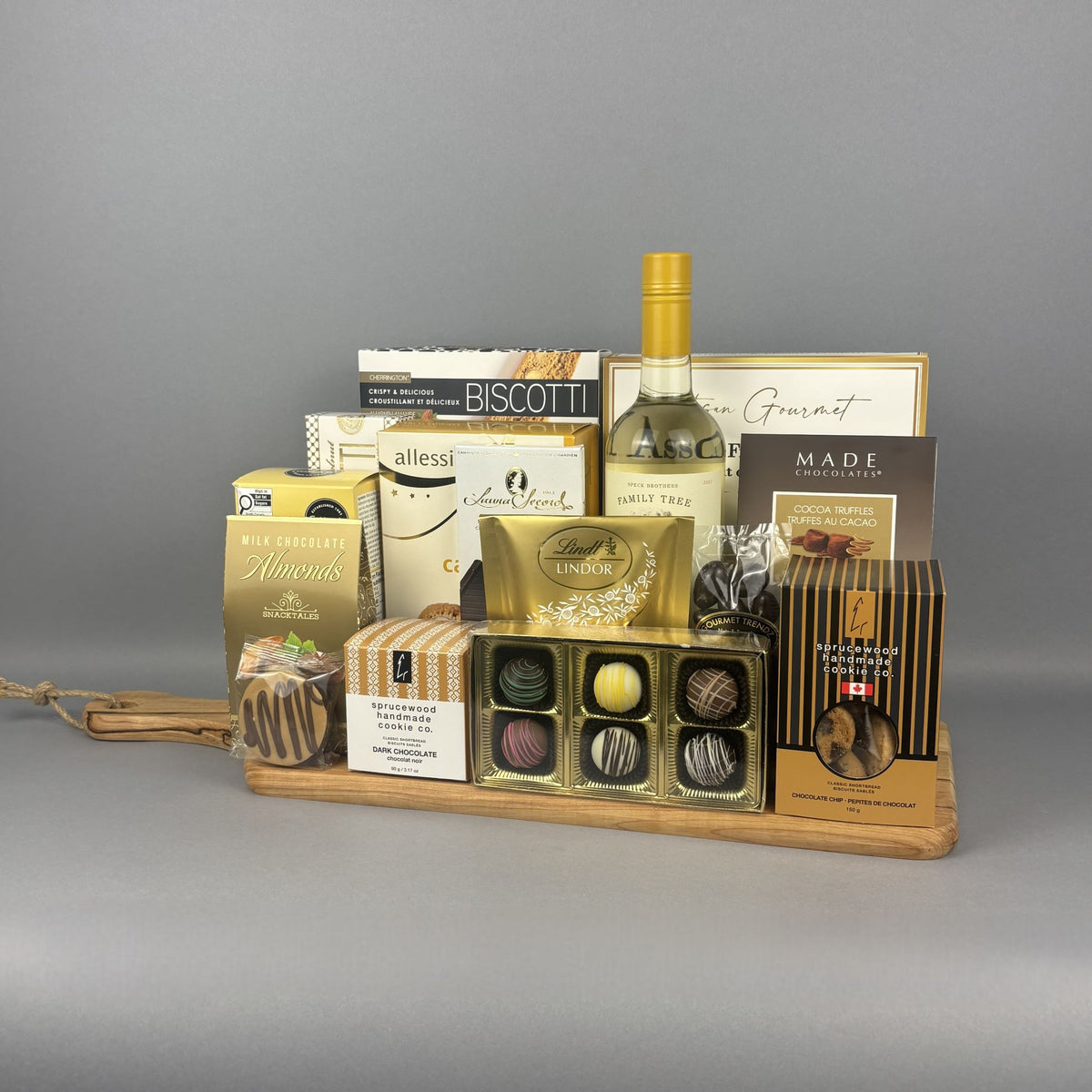 A gourmet gift hamper arranged on a light wood tray. The hamper includes a bottle of white wine, assorted biscotti, chocolates, truffles, and packaged snacks from brands such as Sprucewood, Laura Secord, and Lindt.