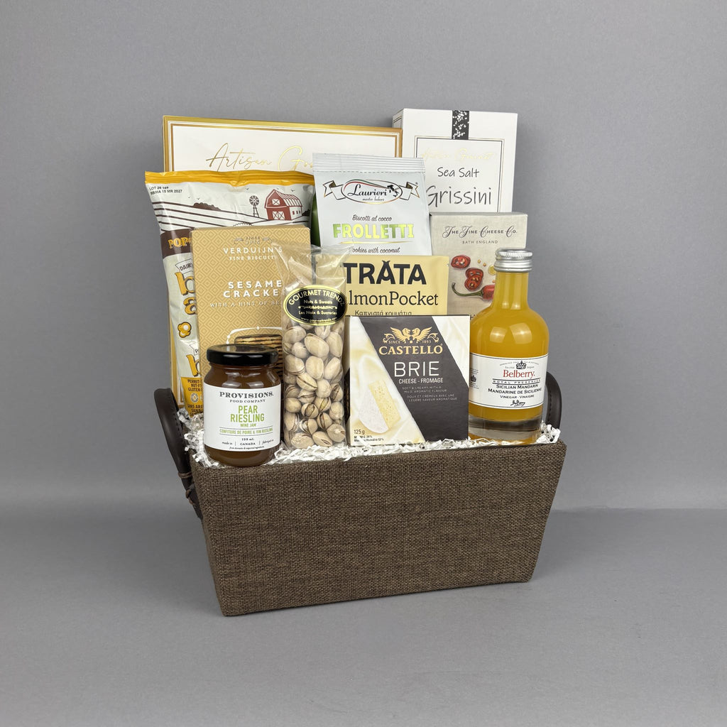 A gourmet gift basket filled with assorted food items including a bottle of mandarin vinegar, Castello Brie cheese, pistachios, sesame crackers, and butter popcorn, arranged in a brown woven basket.