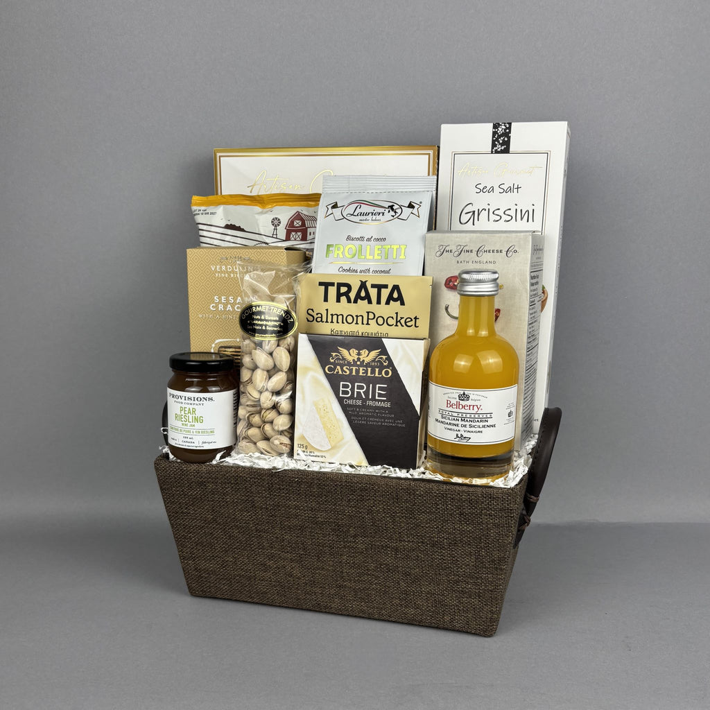 A gourmet gift basket arranged in a brown woven basket, containing a jar of Provision pear riesling jam, a bottle of mandarin vinegar, Castello Brie cheese, froletti coconut cookies, Sea Salt breadsticks, and other packaged snacks and treats.