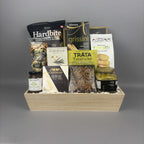 A gourmet gift hamper arranged in a light wood tray, containing packaged snacks and foods including Hardbite chips, breadsticks, Tràta Tuna Pocket, Castello Brie, and a jar of Provisions jam.