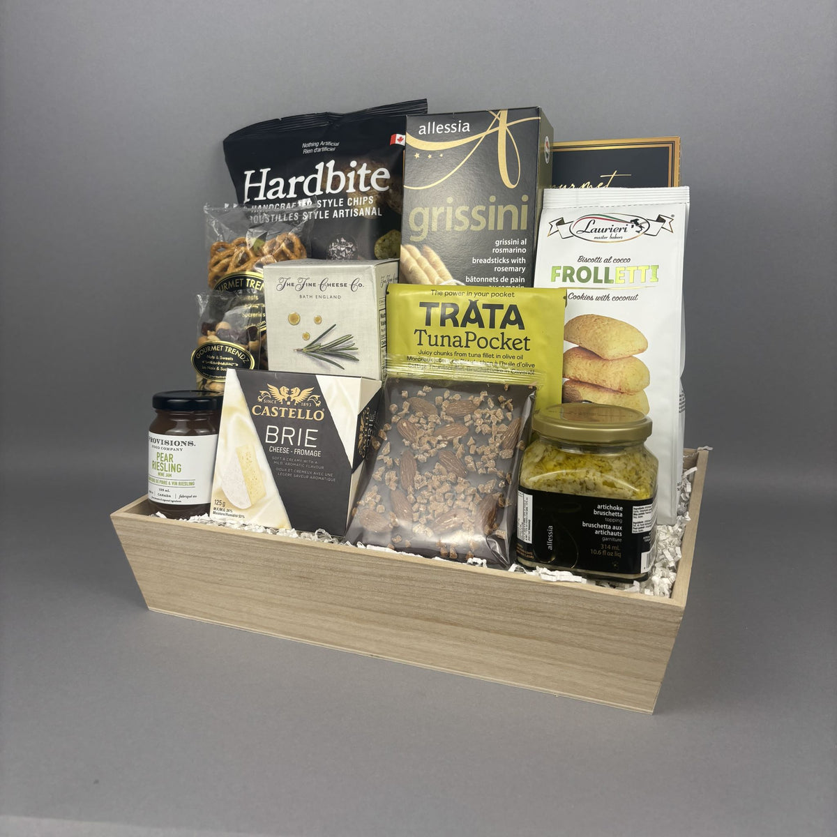 A gourmet food gift basket arranged in a light wood tray, featuring packaged snacks and treats from brands such as Hardbite, Allessia, Provisions, and Castello.