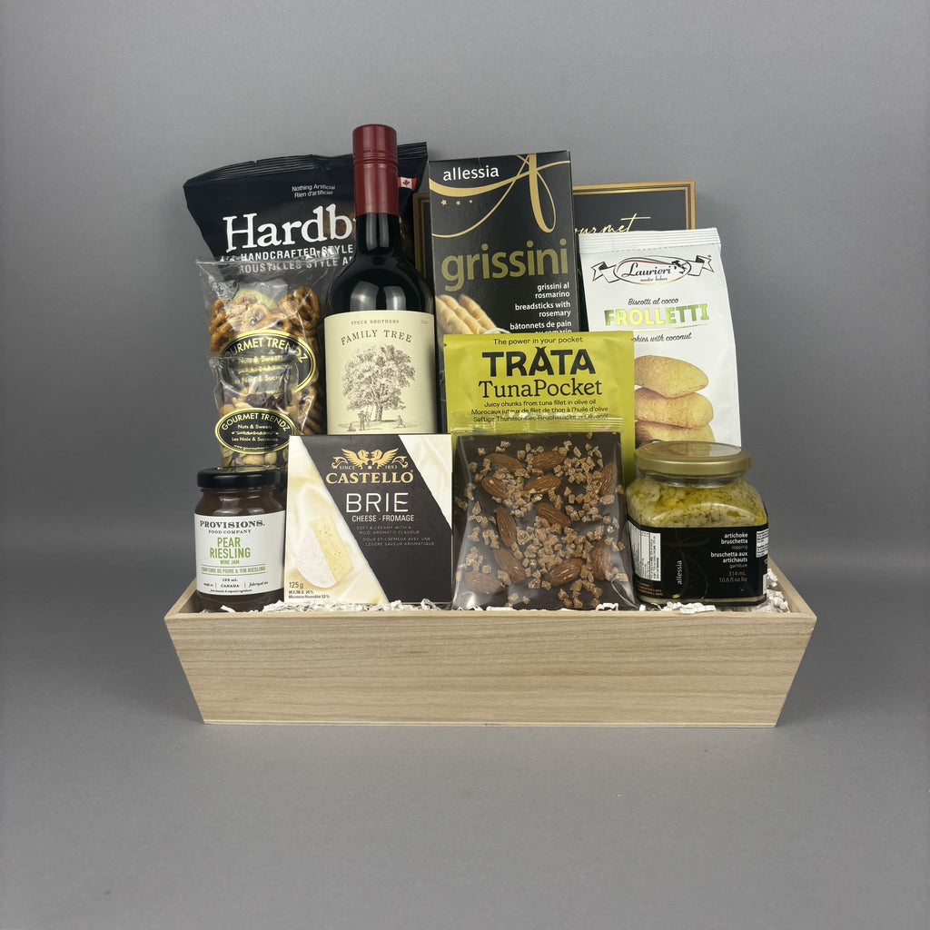 A gourmet gift basket arranged in a light wood tray. The basket contains a bottle of red wine, bags of chips, cookies, jam, and other treats from brands like Hardbite, Provisions, Laurieri, and Castello.