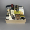 A gourmet gift basket arranged in a light wood tray. The basket contains a bottle of red wine, bags of chips, cookies, jam, and other treats from brands like Hardbite, Provisions, Laurieri, and Castello.