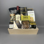 A gourmet gift basket arranged in a light wood tray. The basket contains a bottle of red wine, bags of chips, cookies, jam, and other treats from brands like Hardbite, Provisions, Laurieri, and Castello.
