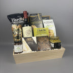 A gourmet gift hamper arranged in a light wood tray, featuring a bottle of red wine, packaged snacks, cheese, and a jar of artichoke bruschetta.