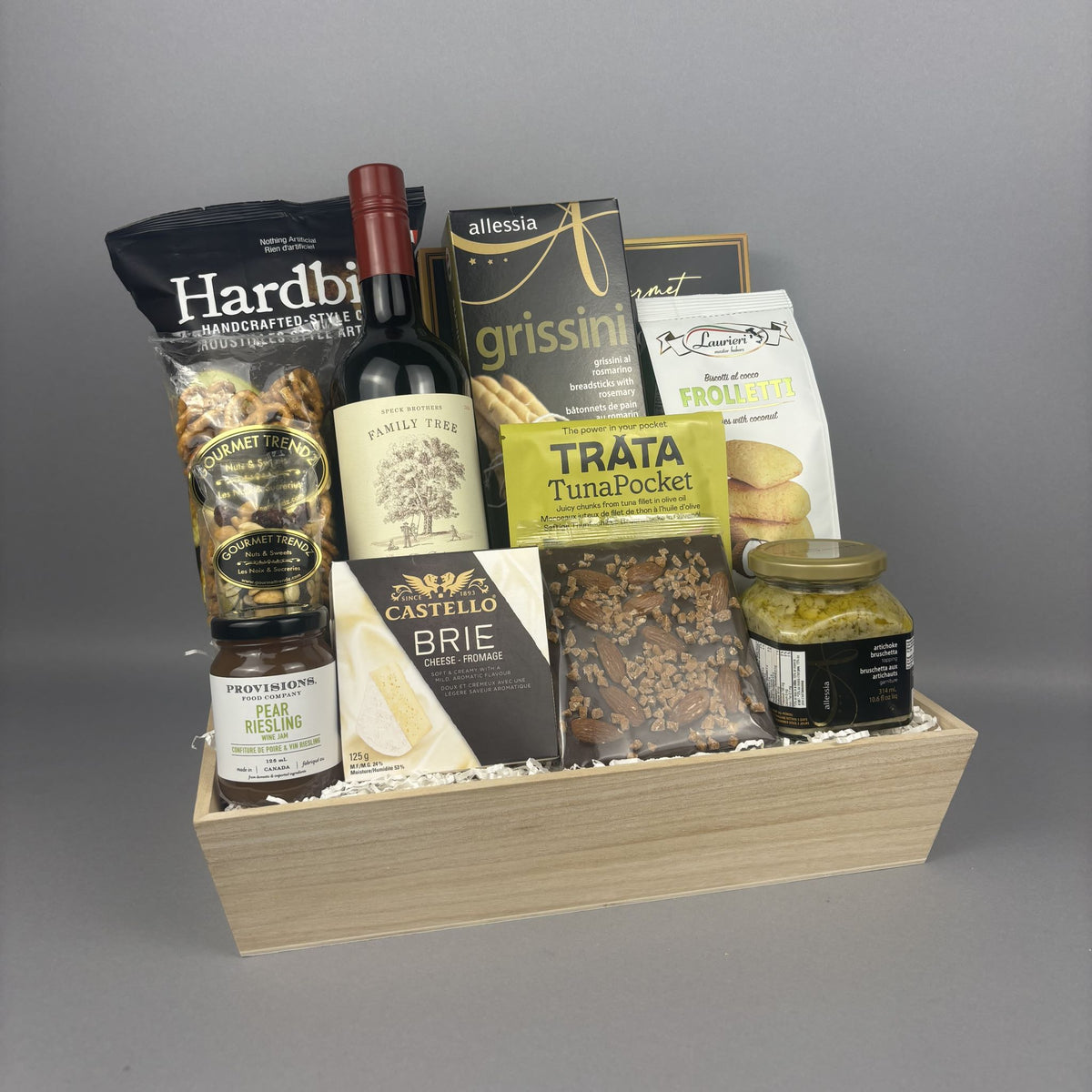 A gourmet gift hamper arranged in a light wood tray, featuring a bottle of red wine, packaged snacks, cheese, and a jar of artichoke bruschetta.