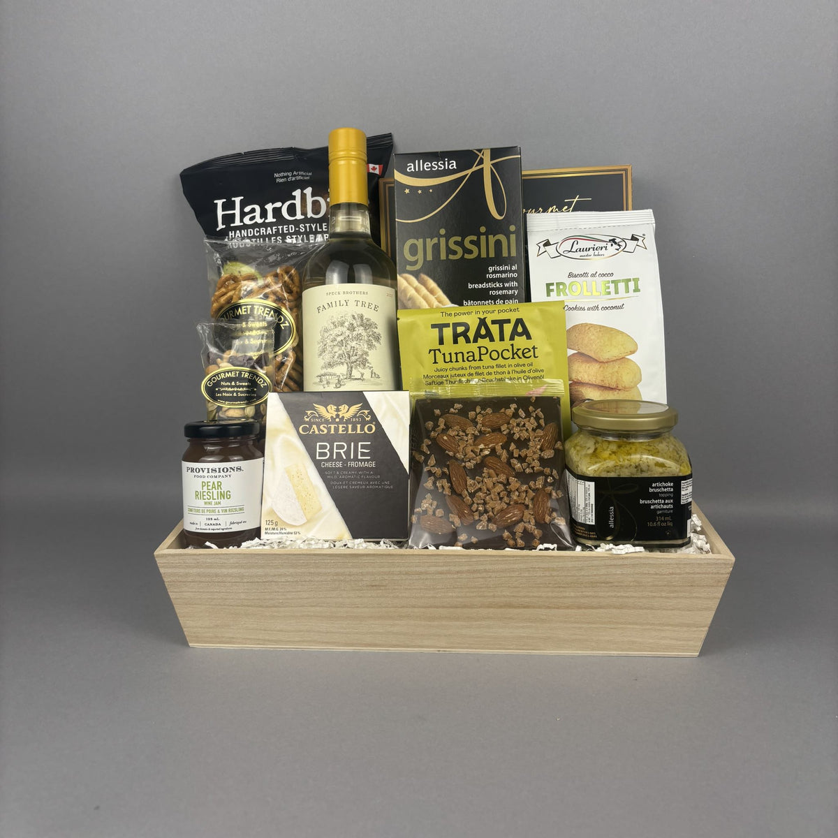 A gourmet gift hamper arranged in a light wood tray, featuring packaged snacks, a bottle of white wine, and jars of jam and artichoke bruschetta. Visible brands include Hardbite, Laurieri, Allessia, TRATA, Castello, and Provisions.