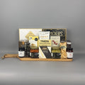 A gourmet gift hamper arranged on a wooden serving board. The set includes a box of Castello Brie Cheese, Salmon Pocket, chili crackers, sesame crackers, jars of preserves and spreads, and shortbread cookies, on a large wooden cutting board.