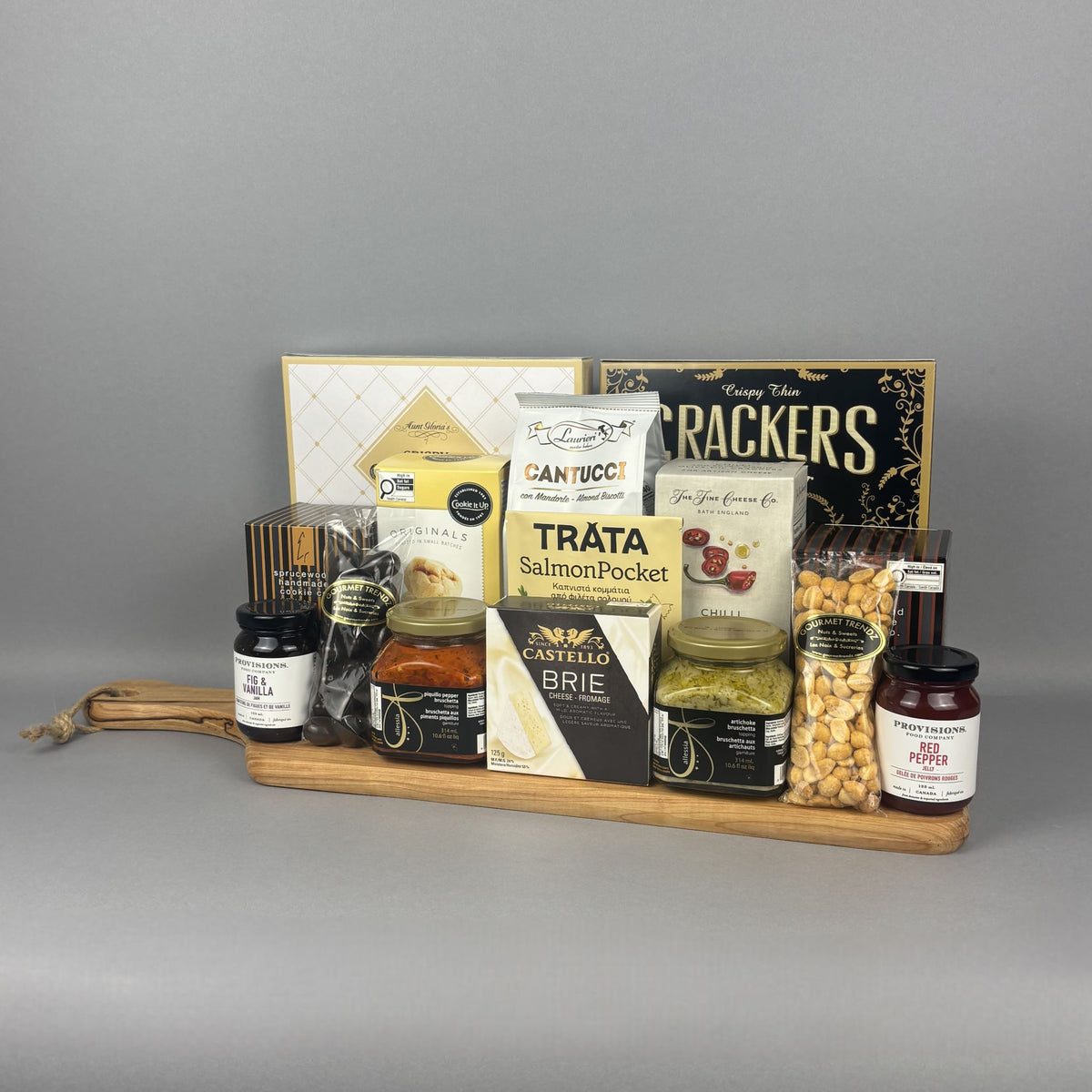 A gourmet gift hamper arranged on a wooden tray. The hamper includes a box of crackers, a package of salmon pate, a box of brie cheese, jars of preserves, a bag of nuts, and shortbread cookies.