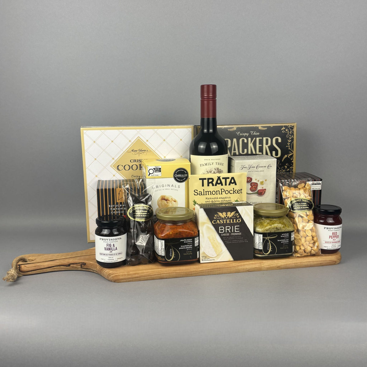 A gourmet gift hamper arranged on a wooden serving board. The hamper includes a bottle of red wine, assorted cheeses, crackers, spreads, and packaged snacks.