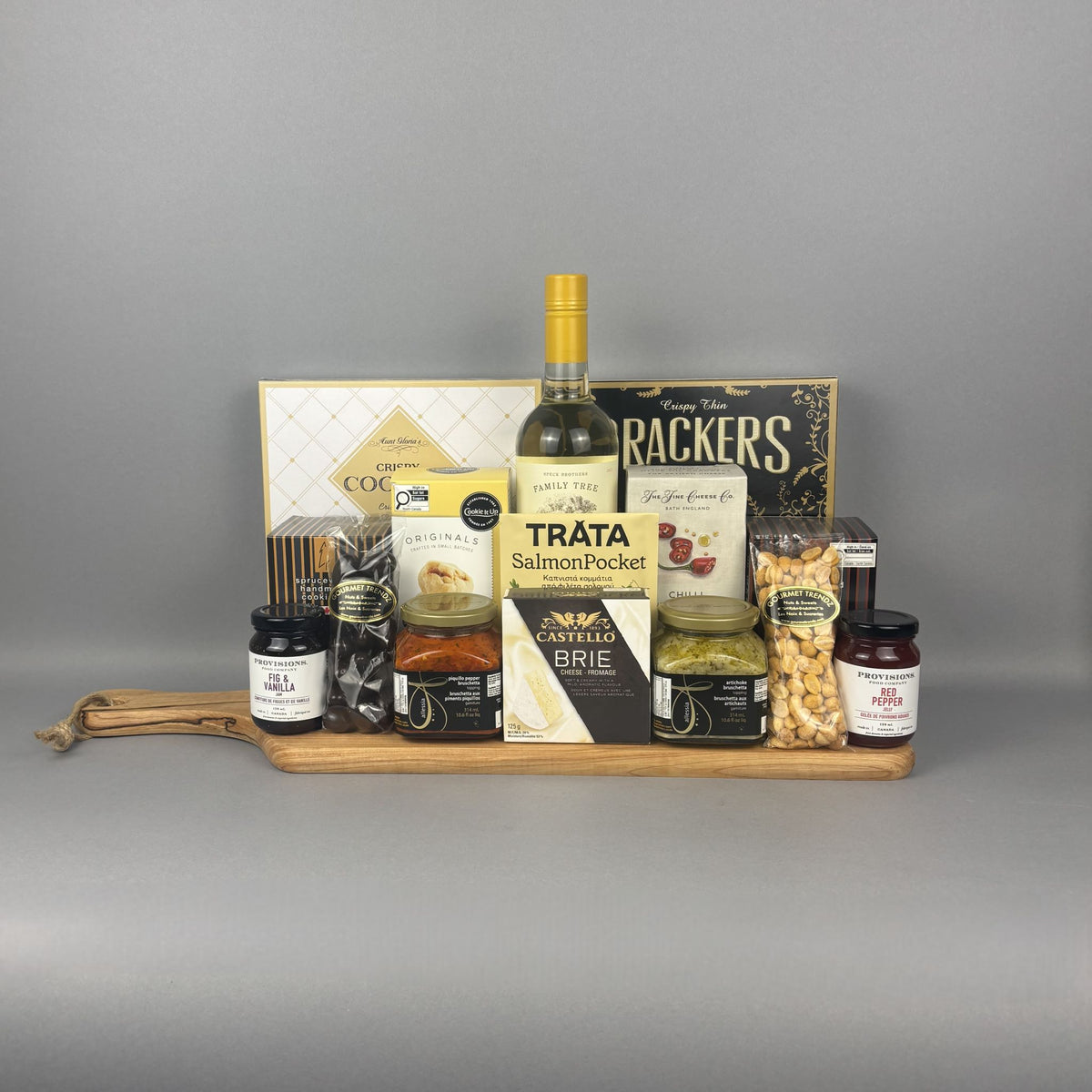 A gourmet gift hamper arranged on a wooden serving board. The hamper includes a bottle of white wine, a box of crackers, a package of smoked salmon, a box of brie cheese, jars of spreads and preserves, and bags of nuts.