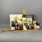 A gourmet gift hamper arranged on a wooden serving board. The hamper includes a bottle of wine, assorted cookies, crackers, jams, spreads, and packaged snacks.