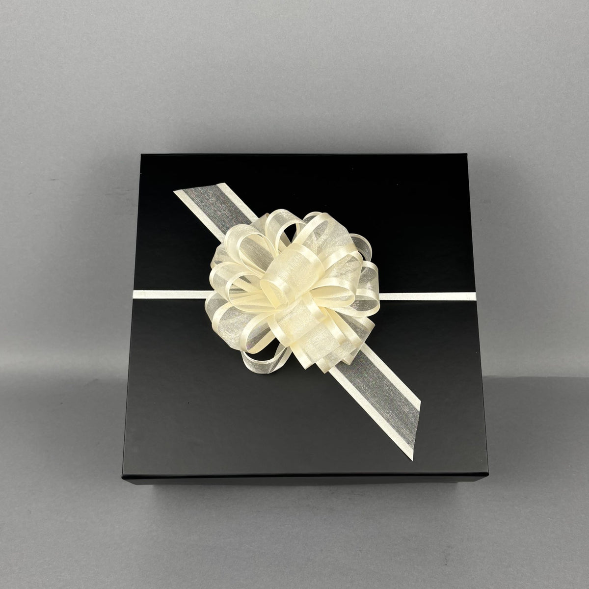 A square black gift box wrapped with a white ribbon and a large cream-colored decorative bow.