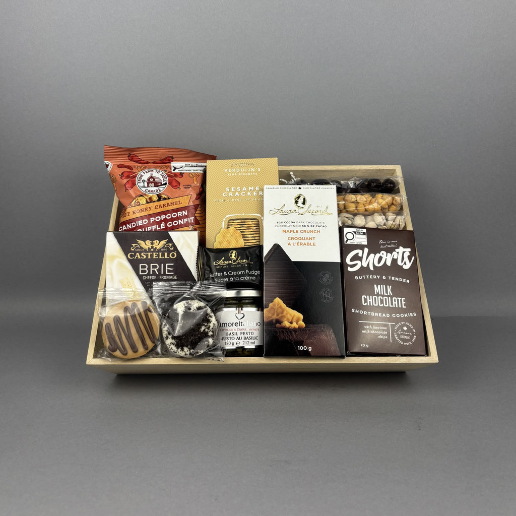 A gourmet chocolate and snack gift tray arranged in a light wood box. The tray includes a bag of Hot Honey Caramel Popcorn, a box of Sesame Crackers jar of basil petso, Laura Secord Maple Crunch Dark chocolate bar, a box of Short's Milk Chocolate Shortbread cookies, a box of Brie cheese, and assorted nuts.