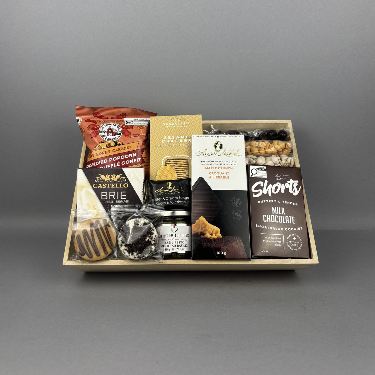 A gourmet chocolate and snack gift tray arranged in a light wood box. The tray includes a bag of Hot Honey Caramel Popcorn, a box of Sesame Crackers jar of basil petso, Laura Secord Maple Crunch Dark chocolate bar, a box of Short's Milk Chocolate Shortbread cookies, a box of Brie cheese, and assorted nuts.