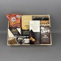 A gourmet snack and chocolate gift box arranged in a light wood tray. The box contains a bag of caramel popcorn and chocolate cover oreos, a box of Sesame Crackers, a box of Castello Brie, a jar of Amore Basil Petso, a box of Short's Milk Chocolate Shortbread cookies, and assorted nuts.