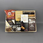 A gourmet snack and chocolate gift box arranged in a light wood tray. The box contains a bag of caramel popcorn and chocolate cover oreos, a box of Sesame Crackers, a box of Castello Brie, a jar of Amore Basil Petso, a box of Short's Milk Chocolate Shortbread cookies, and assorted nuts.