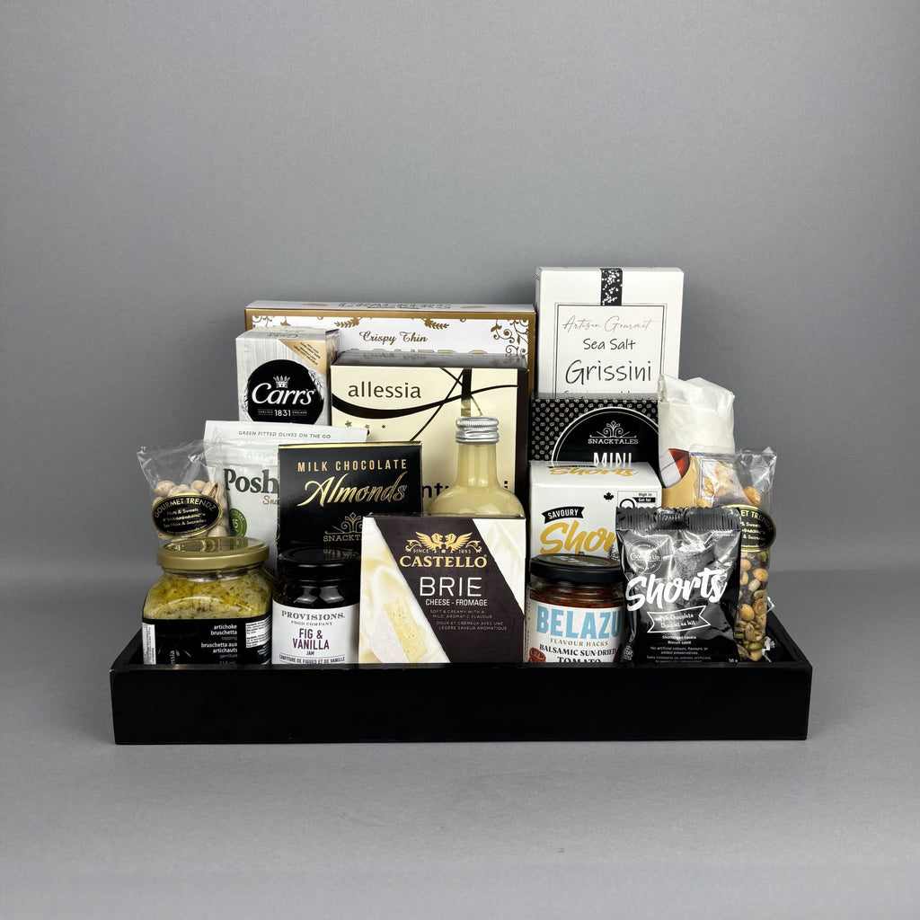 A curated gourmet gift hamper arranged in a black serving tray. The assortment includes cheese, chocolate, nuts, and spreads such as Castello Brie, Allessia artichoke bruschetta, Belazu Balsamic Sundried Tomato, and various crackers and cookies.