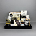 A curated gourmet gift hamper arranged in a black serving tray. The assortment includes cheese, chocolate, nuts, and spreads such as Castello Brie, Allessia artichoke bruschetta, Belazu Balsamic Sundried Tomato, and various crackers and cookies.