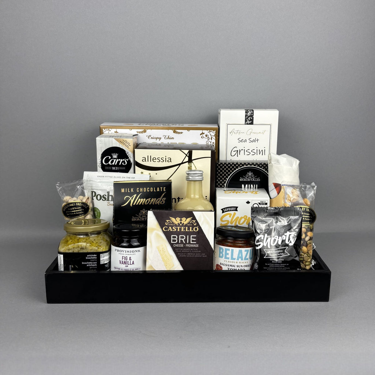 A curated gourmet gift hamper arranged in a black serving tray. The assortment includes cheese, chocolate, nuts, and spreads such as Castello Brie, Allessia artichoke bruschetta, Belazu Balsamic Sundried Tomato, and various crackers and cookies.