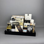 A gourmet gift hamper arranged in a black wooden tray. The tray contains an assortment of packaged food items including Castello Brie cheese, milk chocolate almonds, sea salt breadsticks, and various jars and bags.