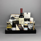 A gourmet gift hamper arranged in a black tray, featuring a bottle of Family Tree red wine, Castello Brie cheese, Belazu tomato spread, sea salt breadsticks, Poshi green olives, Milk Chocolate almonds, and various other gourmet snacks and treats.