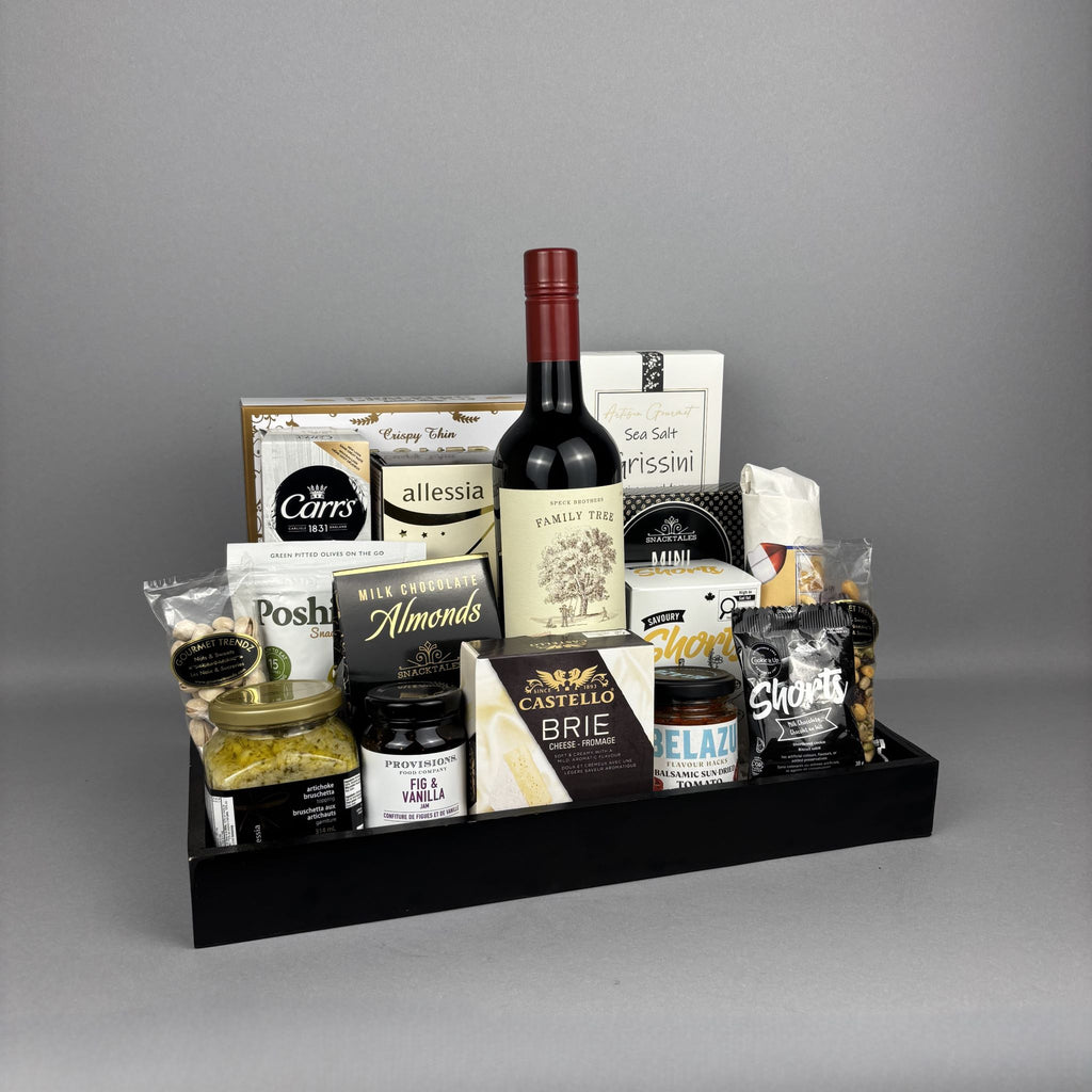 A gourmet gift hamper arranged in a black tray, featuring a bottle of red wine, assorted cheeses, chocolates, nuts, and packaged snacks.